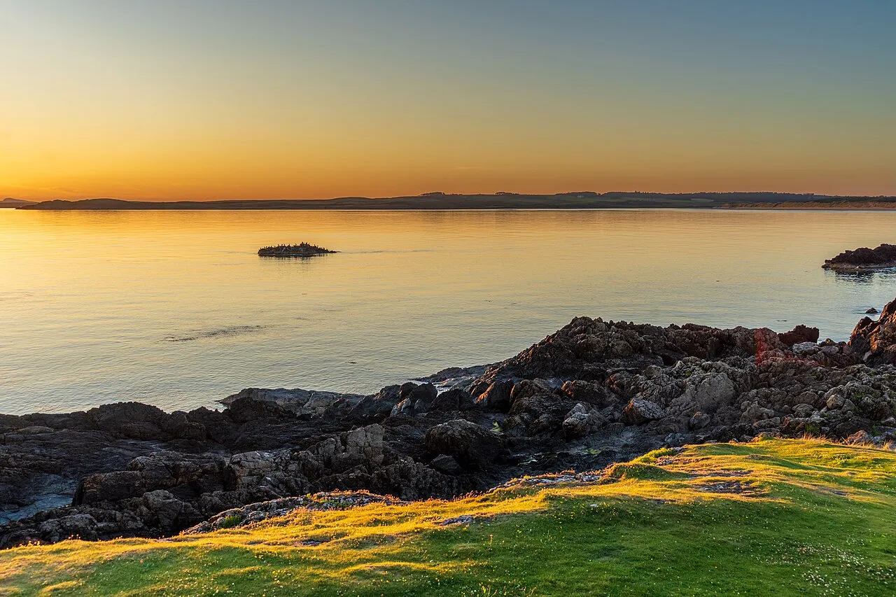 Golden-hour light over a calm sea at Llanddwyn, Anglesey, with dark rocks in the foreground and a low island on the horizon.