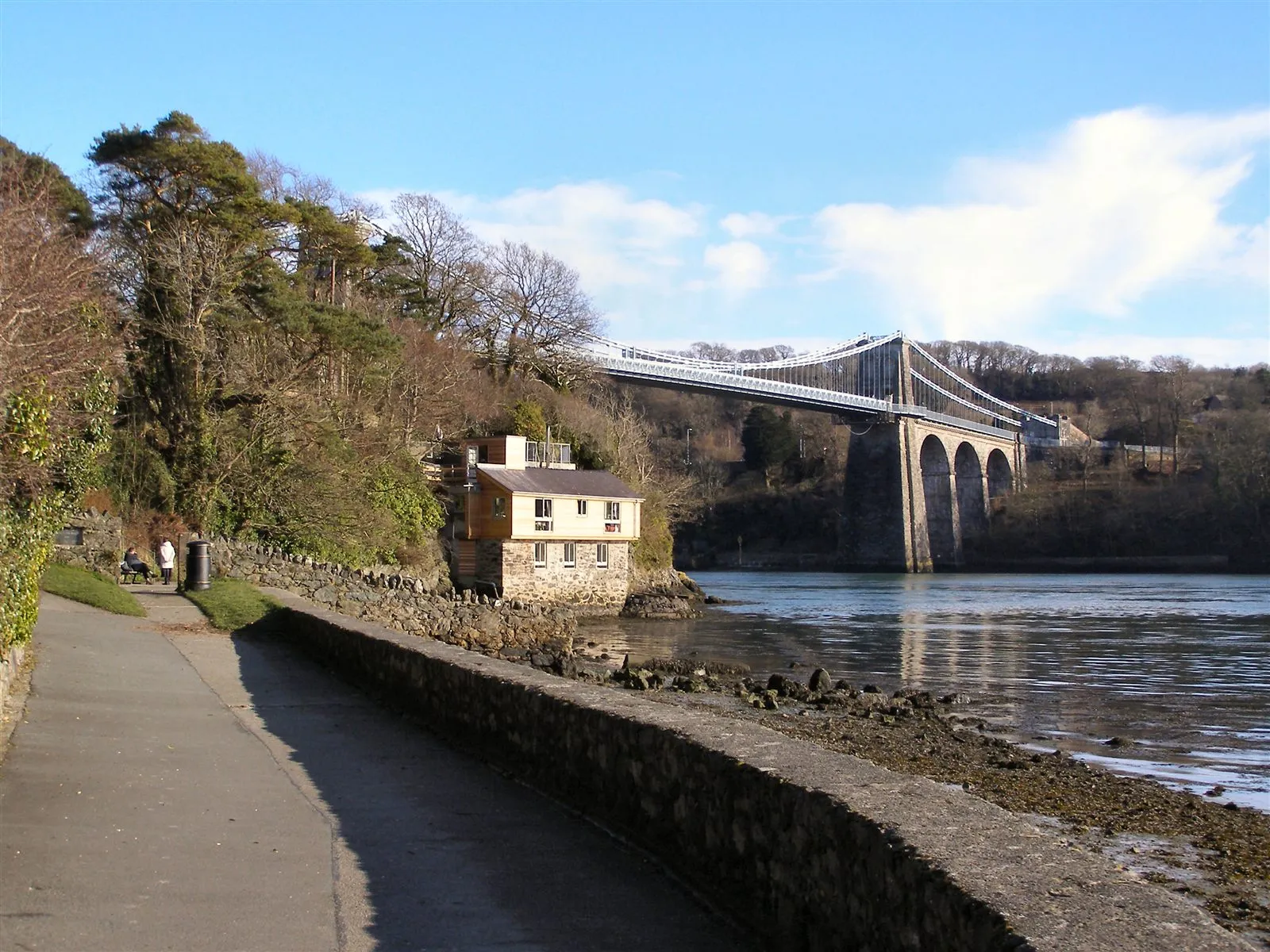 The Belgian Promenade waterside path in Menai Bridge village, with the old pilot's boathouse on the shore and the suspension bridge crossing the strait beyond.