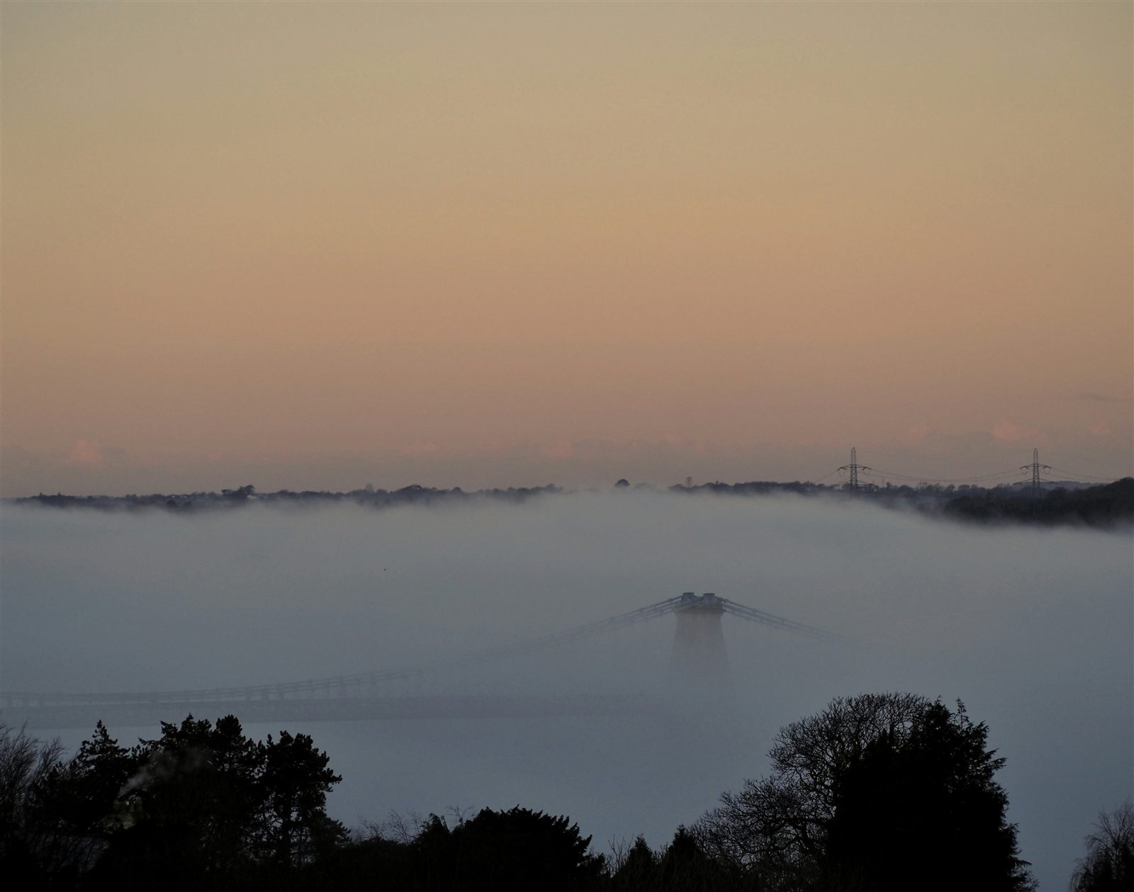 The Menai Suspension Bridge emerging as a faint silhouette through thick coastal fog at sunrise, its towers and chains barely visible above the mist.