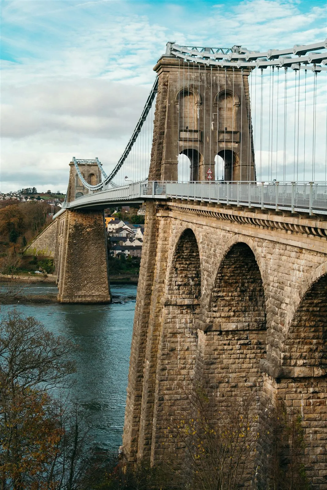 Close view of one of the Menai Suspension Bridge's stone towers, its arched openings and chain anchorages rising above the deck and the limestone viaduct arches below.