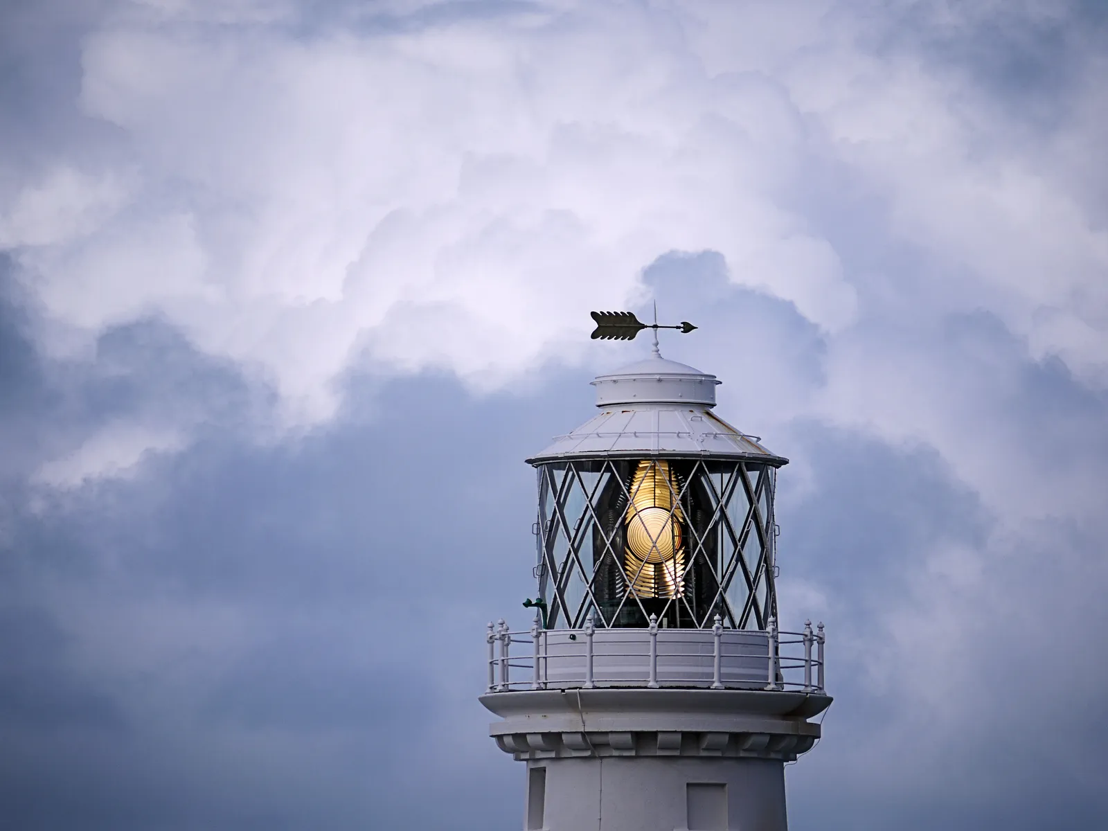 The lit lantern of South Stack Lighthouse close up, the lens glowing behind diamond glazing beneath a weathervane and clouded sky.